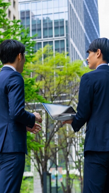 パソコンを持った男性とスーツ姿の男性が屋外で話している様子